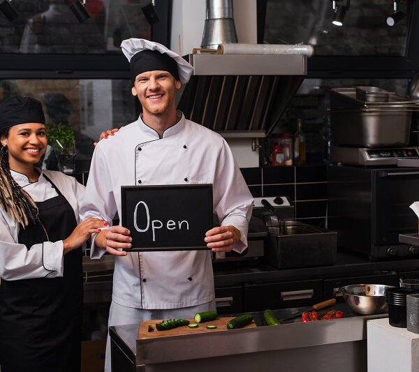 happy young chef in uniform holding chalkboard with open lettering near african american colleague
