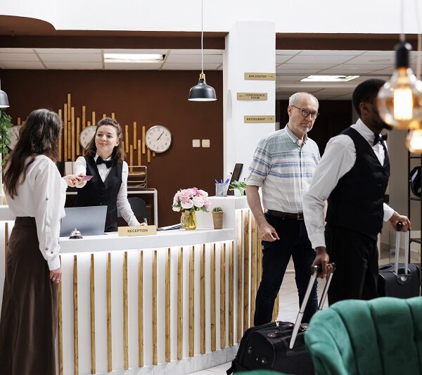 Female concierge assists elderly lady at front desk, providing smooth check-in process and verifying reservations. African american bellboy helps retired senior man with suitcases to hotel room.